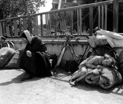 Refugees fleeing the fighting have brought their bicycles and bundles into a school yard. 10th August 1944. A little girl is lying exhausted on a rolled blanket, while her grandmother is sitting next to her. Her white cap is a "bachelique" knitted crochet. Many people from the fighting in Mortaine, just to the south have been seeking refuge in Saint-Pois during August 1944. Mortaine was the final battle for Nornandy. Saint-Pois, Normandy, France. (Photo by Galerie Bilderwelt/Getty Images)